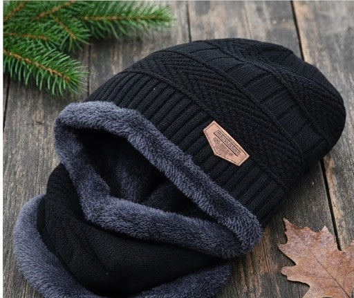 Black knit beanie with gray fleece lining on a wooden surface with a pine branch and leaf.